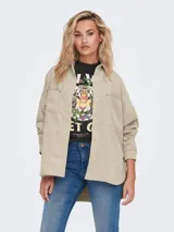 Camisa oversize color beige de pana con cuello clásico, cierre frontal con botones, mangas largas con puños abotonados, dos bolsillos con botón en el pecho y ruedo asimétrico.