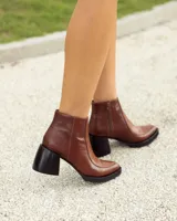 Botín corto de cuero marrón, estilo cowboy moderno, con taco ancho y bajo de color negro. Presenta un panel frontal con textura de piel de reptil y punta cuadrada.