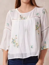 Blusa blanca de gasa con bordados florales, corte holgado, mangas 3/4 acampanadas con detalle de puntilla y forro interno.
