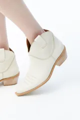Botas de caña baja color crema, estilo texanas, con bordados a tono en la puntera y caña. Cuentan con un tacón bajo y punta alargada redondeada con suela cuadrada de terminación lisa que sobresale levemente.