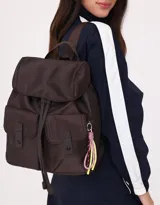 Mochila color crema de tela acolchada con pespuntes verticales. Tiene un bolsillo frontal con cierre, doble cierre superior, manija superior, correas regulables y bolsillos laterales. Incluye un charm colgante de corazón.