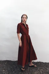 Vestido midi camisero color ladrillo, confeccionado en popelina de algodón. Presenta cuello solapa, manga corta con puños abotonados, bolsillos laterales ocultos y bolsillos de parche con solapa en el pecho. Incluye cinturón lazada en el mismo tejido y cierre frontal con botones ocultos.