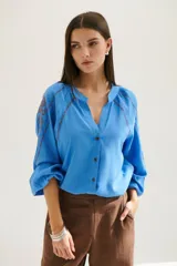 Camisa blanca de manga larga con bordados marrones en hombros y mangas, cuello en V y cierre frontal con botones.