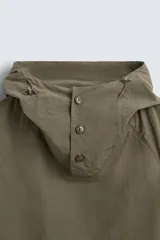 Parka color verde militar de corte amplio, confeccionada en tejido técnico ligero. Cuenta con cuello alto con capucha ajustable, cierre frontal con botones, mangas largas con puños ajustables, bolsillos de plastrón con solapa en la parte delantera, cintura ajustable con cordón y bajo con aberturas laterales ajustables con cordón.