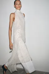 Vestido largo de cuello halter con lazadas ajustables y estampado de lunares negros sobre fondo blanco. Tiene bajo asimétrico, forro interior y cierre con abertura y botón en la espalda.