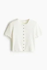 Saco corto color blanco hueso en tejido de punto fino, con cuello redondo, mangas cortas, botonadura frontal y ribete festoneado en tapeta, mangas y bajo.