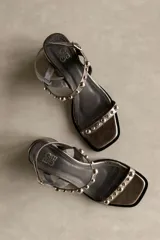 Sandalia de cuero color marrón con taco bloque medio y punta cuadrada. Presenta tachas esféricas doradas en las tiras y hebilla de ajuste en el tobillo.