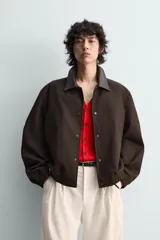 Cazadora de algodón con estructura canvas, color marrón oscuro, de corte relajado. Presenta cuello solapa de cuero negro, cierre frontal con botones a presión, bolsillos de vivo en cadera y puños y bajo elásticos.