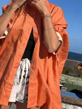 Camisa de lino color naranja durazno, de corte amplio y manga larga con puños anchos que contrastan en color crema/blanco. Presenta cuello camisero y cierre frontal con botones claros.