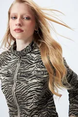 Campera entallada de denim con estampado animal print de cebra en tonos beige y negro. Tiene cuello mao con broches metálicos, cierre frontal con cremallera, bolsillos con cierre en el pecho y mangas largas con puños con cierre.