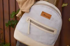 Mochila pequeña de cuero color gris claro y beige, con cierre superior y bolsillo frontal con cierre. Tiene asa de mano y correas ajustables para los hombros.