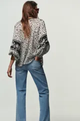 Blusa blanca con estampado floral en negro, cuello redondo con escote pico y manga larga abullonada. Presenta bordados y abalorios en el escote y los puños. Cierre frontal con botones.