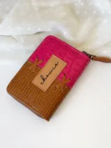 Billetera pequeña de cuero color fucsia y marrón con textura tipo piel de cocodrilo. Tiene cierre metálico y detalle de tiras de cuero cruzadas en el frente.