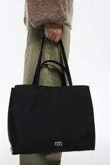 Bolso shopper grande de nylon negro, con estructura blanda, doble asa de mano y correa larga ajustable para llevar al hombro. Presenta un logo metálico con el texto "BIMBA Y LOLA" en la parte frontal inferior.
