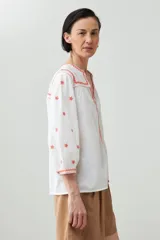 Blusa blanca de corte holgado con bordados de estrellas y detalles geométricos en color coral.