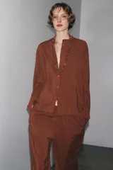 Camisa marrón de Zara Woman Collection confeccionada en hilatura con mezcla de lyocell. Tiene cuello redondo, manga larga acabada en puño, bajo asimétrico con aberturas laterales y cierre frontal con botones.