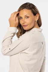 Camisa color beige de lino, de manga larga con botones metálicos personalizados y presilla en la manga. Tiene cuello clásico, cierre frontal parcial con botones y bolsillo de parche en el pecho.