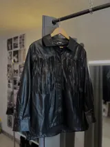 Sobrecamisa negra de ecocuero con flecos en la parte superior delantera y cierre con botones a presión.
