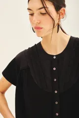 Camisa negra de manga corta con combinación de tejido de punto y plano, detalles de volados y frunces en pechera y espalda, acceso con botones fake nacar y medallita marcaria en espalda.