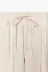 Pantalón de mujer color beige, de corte ancho y tiro medio, confeccionado en tejido fluido. Cuenta con cintura elástica ajustable con cordones, bolsillos laterales y falsos bolsillos de vivo en la espalda.
