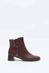 Bota corta de caña baja color marrón oscuro, con puntera cuadrada y taco medio ancho con textura acanalada. Presenta una tira decorativa con hebilla metálica dorada alrededor del tobillo.