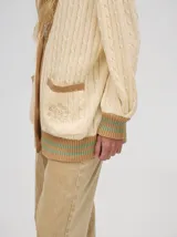 Cardigan largo color crema con detalles en color camel en el borde de los bolsillos, en el borde de los botones y en los puños. Tiene cuello en V y cierre con botones.