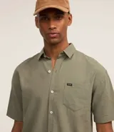 Camisa de hombre de corte relajado, color crudo, con cuello inglés, mangas cortas, abotonado delantero, bolsillo en el pecho y etiqueta de la marca.