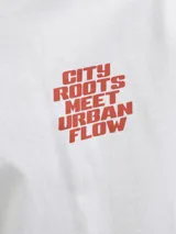 Camiseta blanca de manga corta con cuello redondo, corte holgado y estampado frontal con la frase "City Roots Meet Urban Flow" en color rojo.