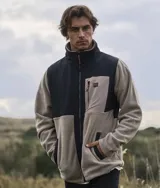 Campera micropolar color beige con bloques en negro y gris. Tiene cierre frontal completo, cuello alto, bolsillos laterales con cierre y logo de la marca bordado en el pecho.