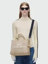 Bolso tote mediano color beige con estampado frontal del logo "The Tote Bag Marc Jacobs" en blanco. Tiene doble asa corta y correa larga extraíble de tela azul marino.