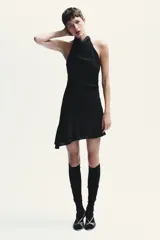 Vestido corto negro de tejido fluido con cuello halter que se ata con lazada. Tiene forro interior, detalle de frunces y bajo asimétrico con volantes. Cierre lateral con cremallera oculta.