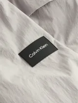Campera Calvin Klein color gris claro, confeccionada en nailon arrugado reciclado, con cuello alto, puños y bajo elásticos, cierre frontal con cremallera, bolsillos laterales con cremallera, abertura de ventilación en la espalda, forro de poliéster reciclado e insignia con el logo de goma en la manga.