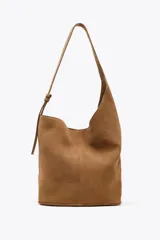 Bolso tipo saca asimétrica color marrón, confeccionado en serraje. Tiene asa bandolera ajustable y cierre con imán. Incluye una cartera con bolsillo extraíble.
