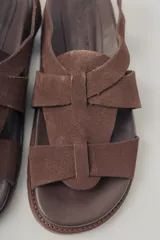 Sandalia plana de gamuza color marrón oscuro, con tiras cruzadas en la parte delantera y una tira fina ajustable con hebilla metálica en el tobillo. Posee plantilla de cuero y suela baja.