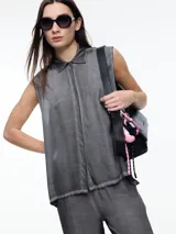 Camisa gris sin mangas confeccionada en viscosa teñida en frío, con efecto gastado. Tiene cuello clásico, cartera oculta y ruedo irregular más largo en los laterales.