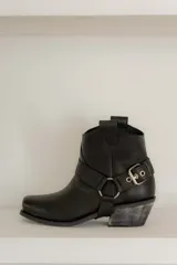 Botas cortas de cuero color marrón oscuro, con puntera cuadrada y caña baja. Presentan una correa con hebilla metálica alrededor del tobillo y un estribo desmontable.