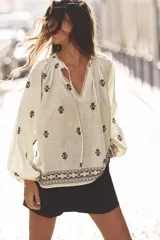 Blusa color crudo de cuello redondo con puntilla, frunces y cordón. Escote pico y manga larga abullonada. Detalle de bordados a contraste en negro.