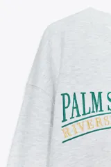 Sudadera gris melange de algodón con cuello redondo, manga larga y texto bordado en verde y azul con la inscripción "Palm Springs USA Riverside County, CA Established 1925".