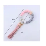 Llavero con pulsera de cuentas redondas de silicona en colores blanco, gris y negro, con una cuenta de madera hexagonal y aro metálico dorado. Incluye borla de flecos de cuero sintético color blanco.
