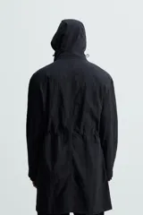 Parka negra de tejido técnico ligero, corte regular, cuello alto con capucha ajustable, manga larga con puños ajustables, cintura ajustable con cordón interno, bolsillos de solapa en la cadera, abertura en la espalda y cierre frontal con cremallera oculta y botones a presión.