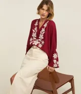 Blusa color borgoña de batista con cuello en V y botones. Presenta bordados florales blancos en el frente y las mangas largas con puños elásticos.