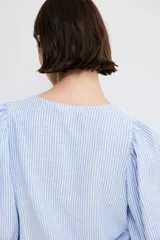 Blusa celeste y blanca a rayas verticales, de corte recto, con cuello redondo, cierre frontal con botones y mangas 3/4 abullonadas con puños elásticos.