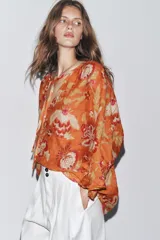 Camisa arrugada de ramio color naranja con estampado floral en tonos rosados y blancos. Tiene cuello redondo con escote pico ajustable con lazadas, mangas 3/4 y bajo asimétrico con aberturas laterales.