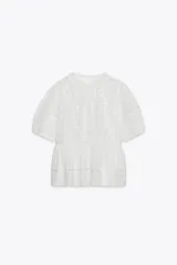 Blusa blanca de algodón con cuello redondo y mangas cortas abullonadas. Presenta bordados florales a tono y cierre con abertura y botón en la espalda.