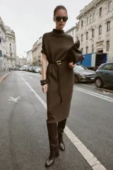 Vestido negro de tejido de punto, corte midi y silueta holgada, con cuello alto acanalado y mangas cortas tipo murciélago. Se ajusta a la cintura con un cinturón fino de efecto piel negro con hebilla dorada en forma de C.