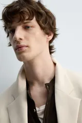 Blazer color beige confeccionado en tejido con lana, de corte relajado. Tiene cuello con solapas de muesca, manga larga con puños abotonados, bolsillo de vivo en el pecho, bolsillos de solapa en la cadera, bolsillo interior, bajo con abertura en el centro de la espalda y cierre frontal con botones.