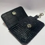 Tarjetero pequeño negro con textura de cocodrilo, cierre con broche metálico y gancho metálico para colgar.