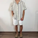 Camisa de manga corta, cuello cubano, color crudo con rayas verticales finas en gris oscuro. Presenta un bolsillo de parche en el pecho y botones marrones.