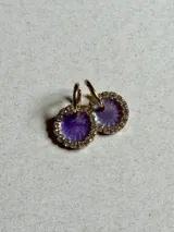 Aros colgantes con dije circular de resina color violeta y borde de strass dorados. El arito es de acero quirúrgico dorado.