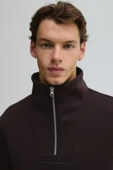 Buzo de tejido pesado color marrón oscuro, con interior cepillado suave. Presenta cuello elevado, medio cierre metálico y hombros caídos. Puños y bajo en punto acanalado.